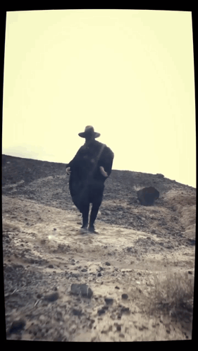 a man wearing a hat and a poncho walks across a dirt road