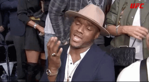 A Man Wearing A Hat And A Ring Is Sitting In A Crowd Of People . GIF