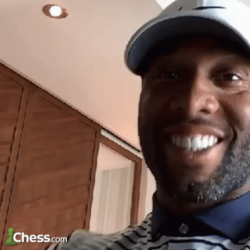A Man Wearing A Hat And A Striped Shirt Is Smiling In Front Of A Sign That Says Chess.com GIF