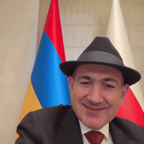 A Man Wearing A Hat And A Suit Smiles In Front Of A Flag GIF