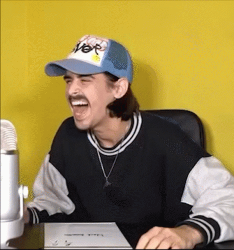 A Man Wearing A Hat And A Sweater Is Sitting At A Desk Laughing . GIF