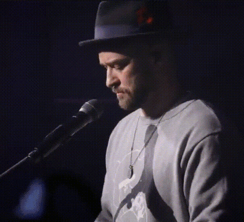 A Man Wearing A Hat And A Sweater Singing Into A Microphone GIF