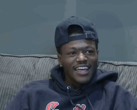 A Man Wearing A Hat And A Sweatshirt With The Letter C On It Sits On A Couch GIF
