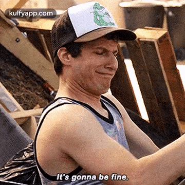 A Man Wearing A Hat And A Tank Top Is Sitting In Front Of A Pile Of Wooden Crates . GIF