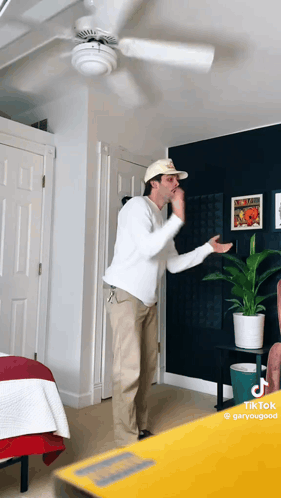A Man Wearing A Hat And A White Shirt Is Standing In A Room With A Ceiling Fan GIF