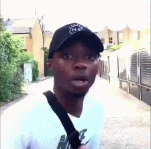 A Man Wearing A Hat And A White Shirt Is Standing On A Sidewalk . GIF