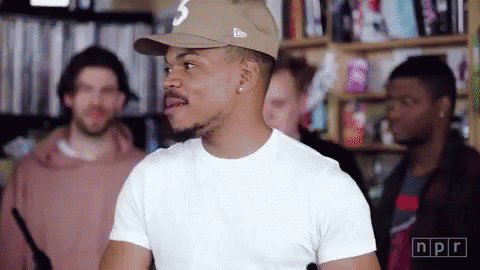 A Man Wearing A Hat And A White T-Shirt Is Standing In A Room With Other People . GIF