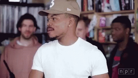 A Man Wearing A Hat And A White T-Shirt Is Standing In Front Of A Group Of Men . GIF