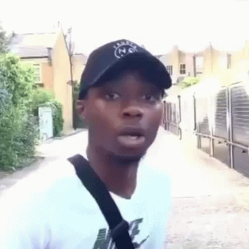 A Man Wearing A Hat And A White T-Shirt Is Standing On A Sidewalk . GIF