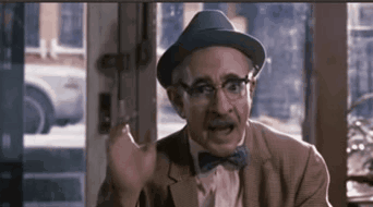 A Man Wearing A Hat And Bow Tie Is Sitting In Front Of A Window And Making A Funny Face . GIF