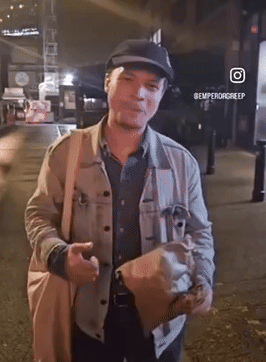 A Man Wearing A Hat And Carrying A Bag Is Standing On A Street With The Hashtag @Emperorgreep GIF
