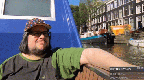 A Man Wearing A Hat And Glasses Is Leaning On A Railing Next To A Body Of Water With Buildings In The Background GIF