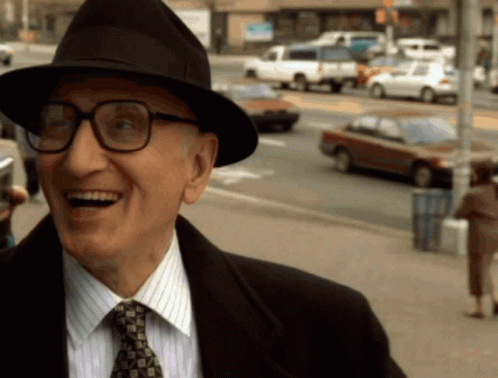 A Man Wearing A Hat And Glasses Is Smiling On The Street GIF
