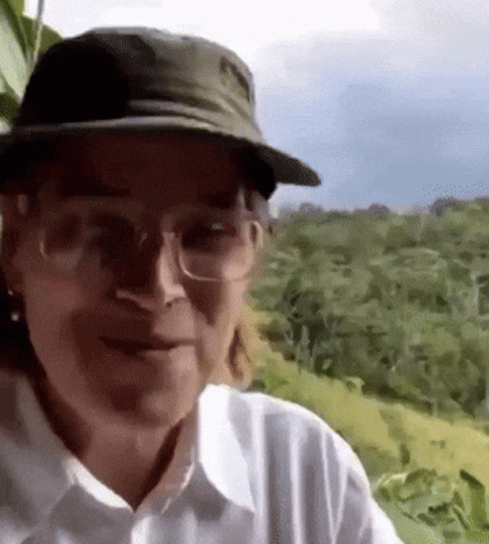 A Man Wearing A Hat And Glasses Is Standing In A Field . GIF