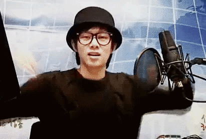 A Man Wearing A Hat And Glasses Is Standing In Front Of A Microphone And Talking Into It . GIF