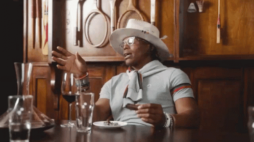A Man Wearing A Hat And Glasses Sits At A Table With Wine Glasses And A Plate Of Food GIF