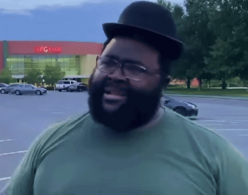 A Man Wearing A Hat And Glasses Stands In A Parking Lot In Front Of A Building That Says The G Rite GIF