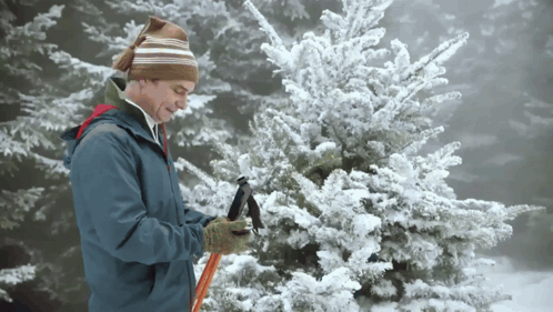 A Man Wearing A Hat And Gloves Is Holding A Hammer In Front Of A Snow Covered Tree GIF