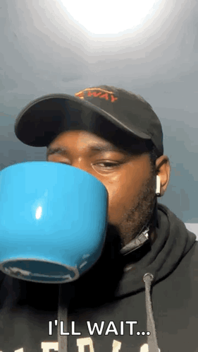 A Man Wearing A Hat And Headphones Is Drinking From A Blue Cup And Saying I 'Ll Wait . GIF