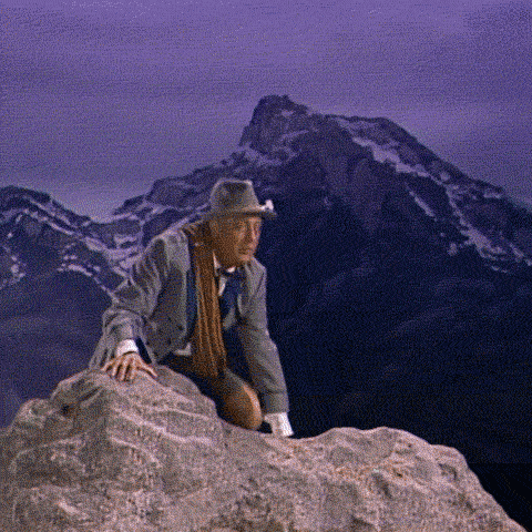 A Man Wearing A Hat And Scarf Is Sitting On A Rock In Front Of A Mountain GIF