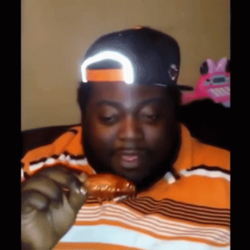 A Man Wearing A Hat And Striped Shirt Is Eating A Chicken Wing GIF