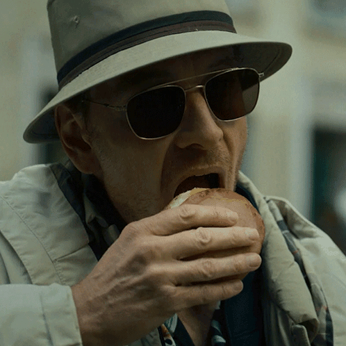 A Man Wearing A Hat And Sunglasses Eats A Sandwich GIF