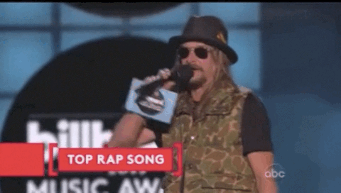 A Man Wearing A Hat And Sunglasses Holds A Microphone In Front Of A Sign That Says " Top Rap Song " GIF