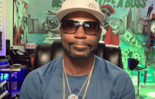 A Man Wearing A Hat And Sunglasses Is Sitting In Front Of A Sign That Says " On Air " GIF