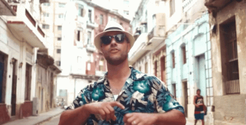 A Man Wearing A Hat And Sunglasses Is Standing On A City Street GIF