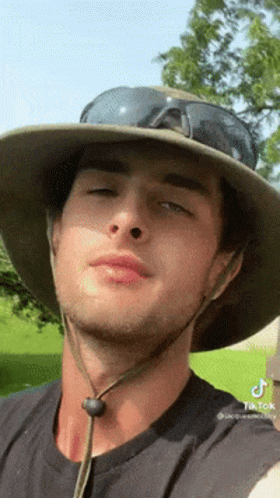 A Man Wearing A Hat And Sunglasses Looks At The Camera . GIF
