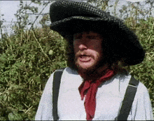 A Man Wearing A Hat And Suspenders Is Standing In Front Of A Bush GIF