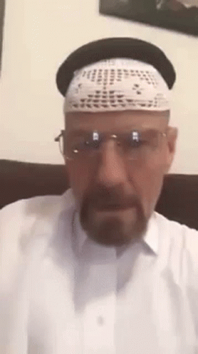 A Man Wearing A Hat , Glasses And A White Shirt Is Sitting On A Couch . GIF