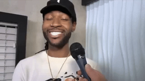 A Man Wearing A Hat Is Holding A Microphone In His Hand And Smiling . GIF
