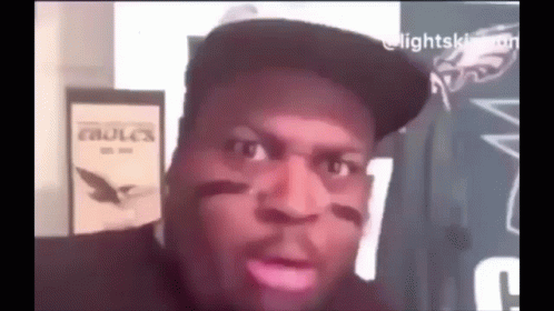 A Man Wearing A Hat Is Making A Funny Face While Sitting In Front Of A Sign That Says Eagles . GIF