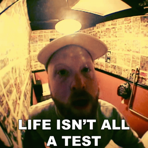 A Man Wearing A Hat Is Sitting In A Bathroom With The Words Life Is N't All A Test Above Him GIF