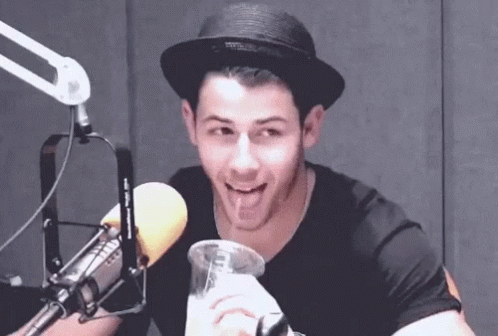 A Man Wearing A Hat Is Sitting In Front Of A Microphone And Drinking From A Glass . GIF
