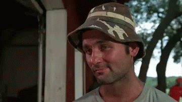 A Man Wearing A Hat Is Standing In Front Of A Building . GIF