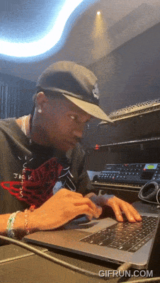 A Man Wearing A Hat Is Typing On A Laptop With Gifrun.com Written On The Bottom Right GIF