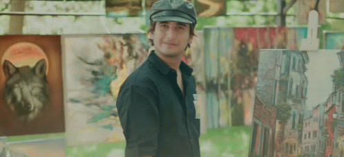 A Man Wearing A Hat Stands In Front Of A Wall Of Paintings Including One Of A Wolf GIF