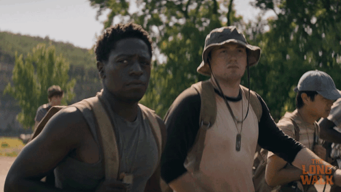 A Man Wearing A Hat Stands Next To Another Man In A Long Walk Advertisement GIF