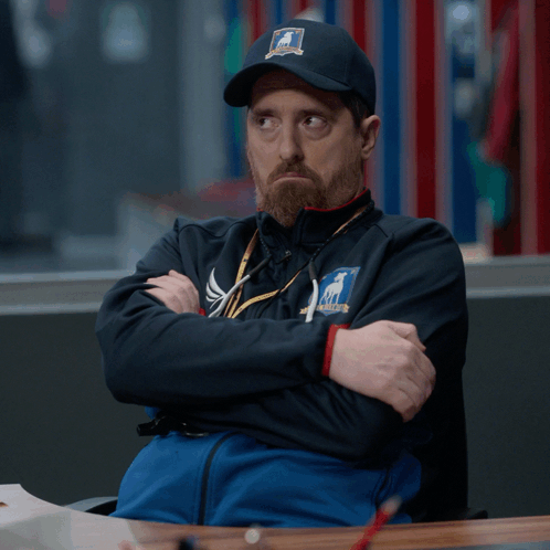 A Man Wearing A Hat That Says ' Chicago ' On It Is Sitting At A Table With His Arms Crossed GIF