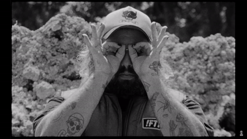 A Man Wearing A Hat With A Skull On It Is Covering His Eyes GIF