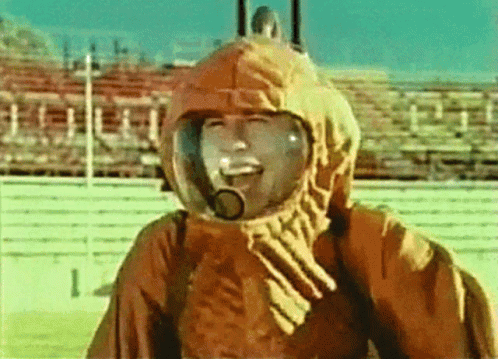 A Man Wearing A Helmet And A Hood Is Smiling In Front Of A Stadium GIF