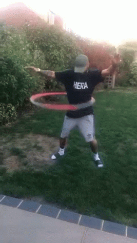 A Man Wearing A Hera Shirt Is Playing With A Hula Hoop In A Yard . GIF