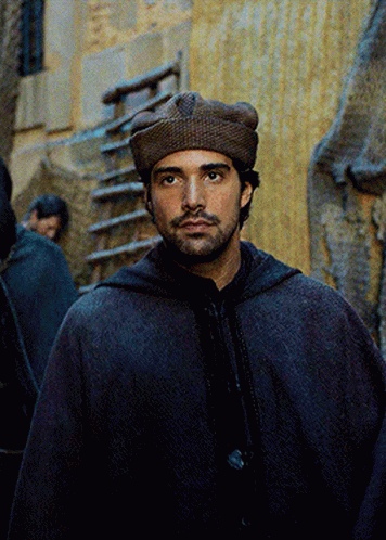 A Man Wearing A Hooded Cape And A Hat Looks At The Camera GIF