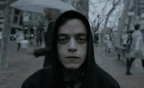 A Man Wearing A Hooded Jacket And Tie Is Standing On A Sidewalk In The Rain . GIF