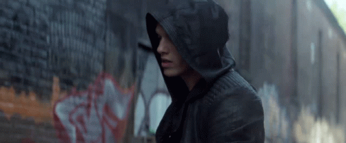A Man Wearing A Hooded Jacket Is Standing In Front Of A Brick Wall With Graffiti On It GIF