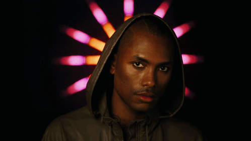 A Man Wearing A Hooded Jacket Stands In Front Of A Pink And Orange Light GIF