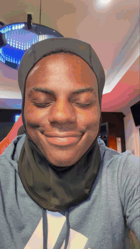 a man wearing a hooded sweatshirt and a scarf around his neck smiles with his eyes closed