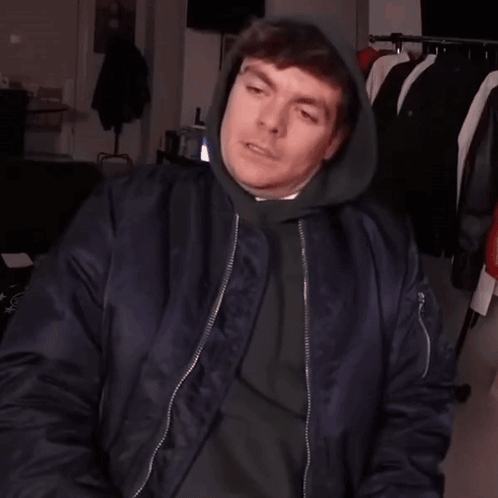 A Man Wearing A Hoodie And A Bomber Jacket Is Making A Hand Gesture GIF